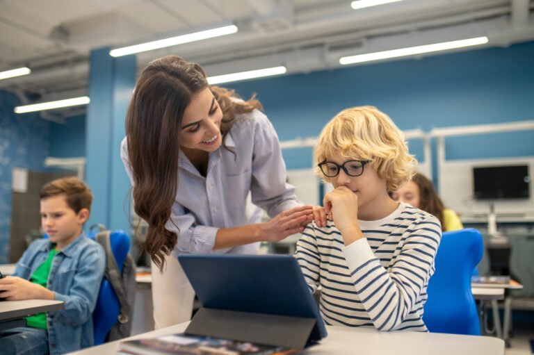 smiling teacher touching shoulder of boy looking at tablet